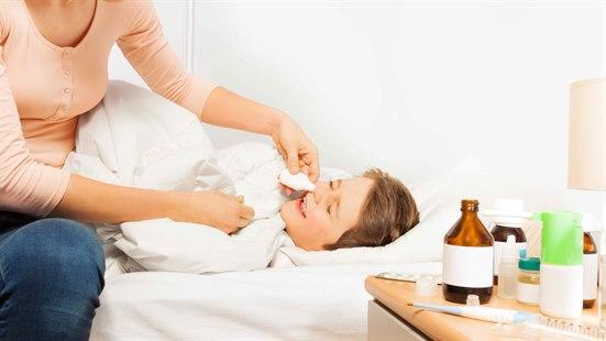 En gutt ligger i sengen, med medisiner og termometer på nattbordet. En damen gir gutten nesedråper.