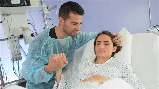 Fødende kvinne ligger i en sykehusseng. Hun holder en mann med blå beskyttelseskjorte i hånden.