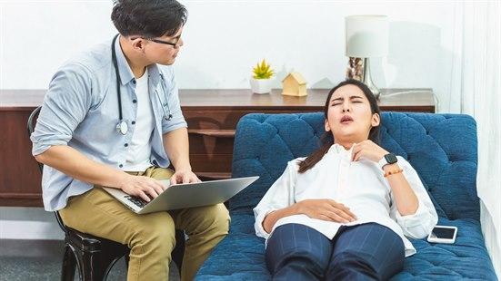 Illustrasjonsfoto: Ung dame ligger på behandlingssofa med lukkede øyne og anstrengt uttrykk. Ved siden av sitter en mann med stetoskop rundt nakken og tar notater på PC-en.