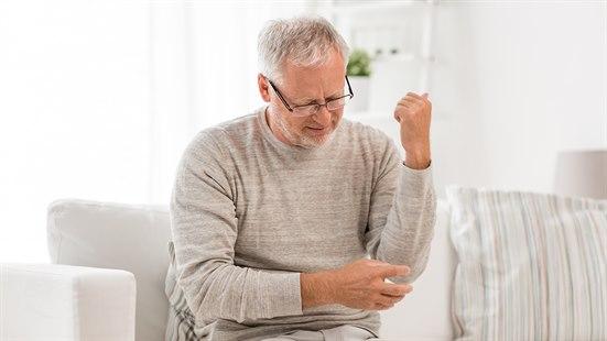 Illustrasjonsfoto av en eldre mann som sitter i en lys stue på en sofa. Han holder den ene hånden på albuen og uttrykker smerte.