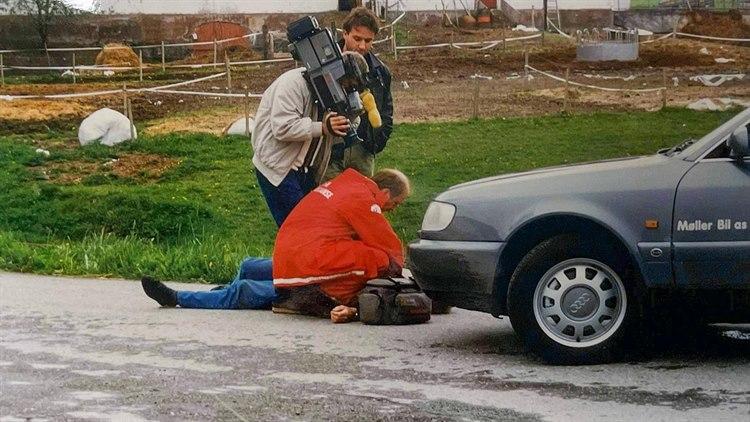 Foto: Mann i rød ambulansedrakt sitter bøyd over en person som ligger på veien, foran en bil. Bak han står to andre menn, den ene med et profesjonelt videokamera.