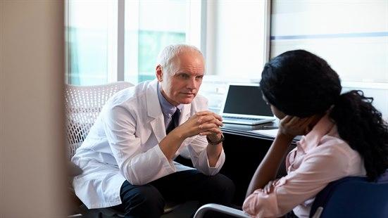 Alvorlig mannlig lege som snakker med kvinnelig pasient. Kvinnen sitter med ryggen mot kamera.