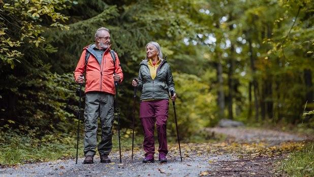 På en skogsti går et par i pensjonsalderen, utstyrt med turlær og staver. De ser ut til å ha det fint mens de går tur og nyter naturen rundt seg.