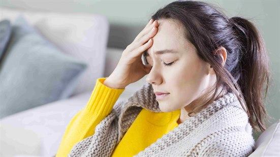 Illustrasjonsfoto: Ung kvinne med gul genser og beige cardigan sitter i sofaen og holder seg for pannen.