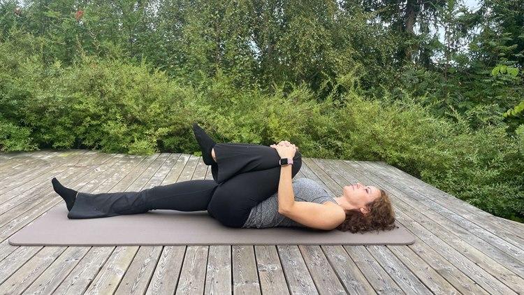 Dame som ligger på ryggen på yogamatte. Utendørs, med trær og busker i bakgrunnen. Ligger flatt på ryggen, med det ene kneet opp mot brystet.
