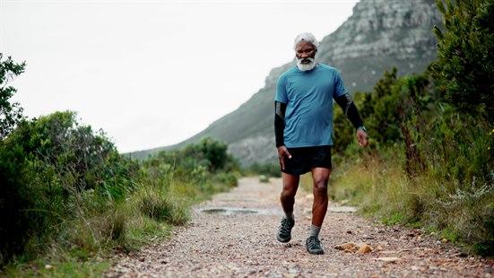 Fotoet viser en eldre svart mann ikledd treningsklær som går tur i et fjellområde. Han tar på sitt høyre lår med sin høyre hånd som om han har vondt. 