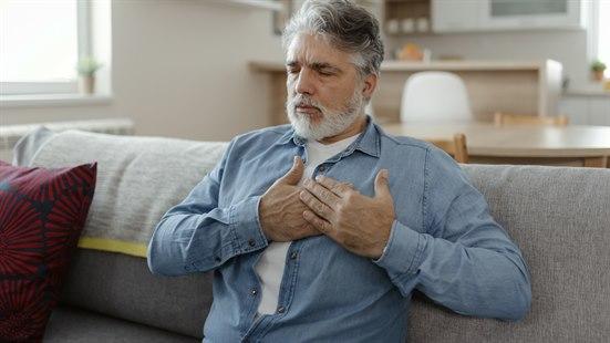 Illustrasjonsfotoet viser et voksen mann som sitter i sofan sin hjemme. Han har hendene over hjertet, og ser bekymret ut.