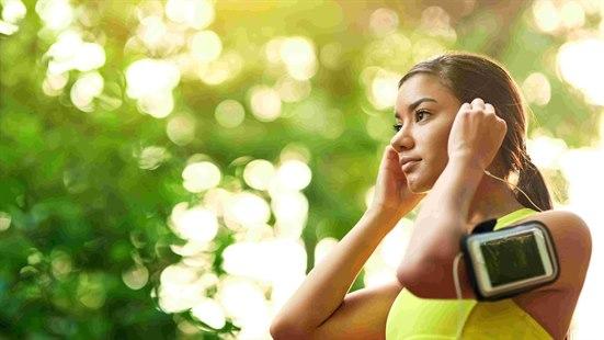 Illustrasjonsfoto: Ung kvinne i gul singlett setter på seg ørepropper. Har smarttelefon i etui som er festet til armen.