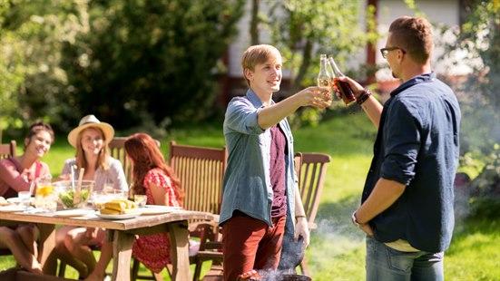 To menn står i hagen og skåler med hver sin flaske. Det er sommer, hageselskp og fint vær. 