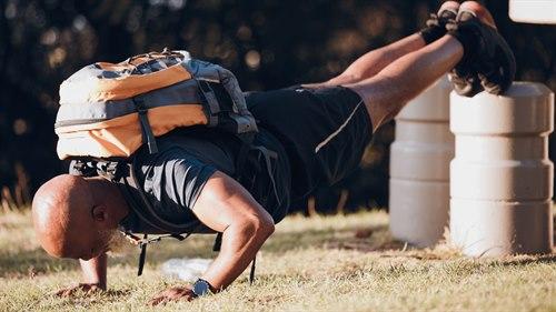 Illustrasjonsfoto: Mann med sort t-skjorte, sorte shorts og oransje ryggsekk gjør skrå armhevinger med beina på noen betongblokker og hendene i bakken.