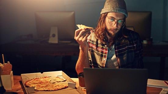 Illustrasjonsfoto: Mann med grå lue, briller og skulderlangs brunt hår spiser pizza foran en laptop i et mørkt kontorlokale. Ved siden av laptopen ligger resten av pizzaen i en eske.