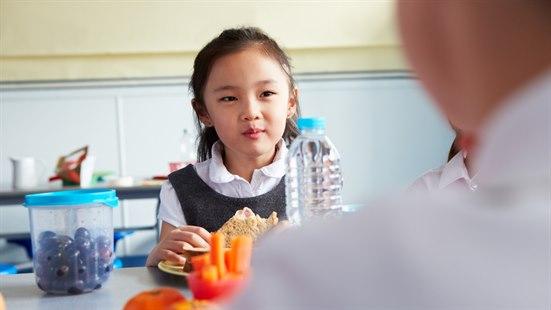 Illustrasjonsfoto: Jente i barneskolealder spiser en brødskive i en kantine. På bordet foran henne ser vi en boks med bær, og en skål med gulrøtter. I front ses litt av bakhodet og skulderen på en annen person.