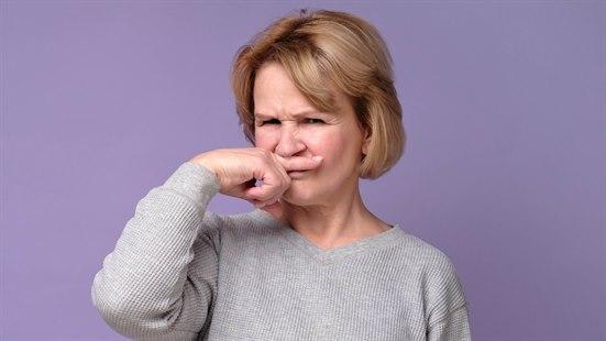 Fotoet viser en kvinne i 60-årene som tar seg i ansiktet mellom nesen og munnen med sin høyre pekefinger. Bildeutsnittet viser den øvre tredjedel av kvinnen, som er ikledd grå genser, foran fiolett bakgrunn. Ansiktet uttrykker ubehag (dårlig lukt).