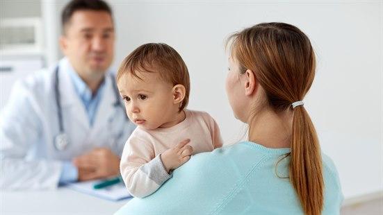 Illustrasjonsfotoet viser en kvinne med et barn på armen. Kvinnen befinner seg på et legekontor der en mannlig lege vises i bakgrunnen