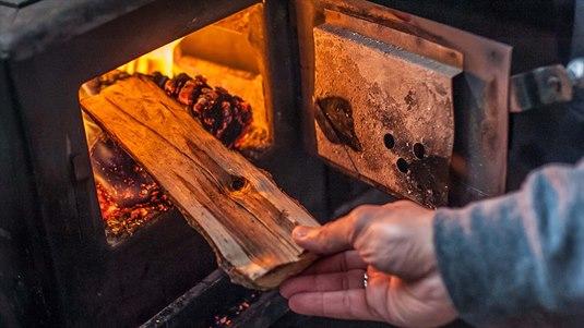 En person legger en vedkubbe inn i et fyringsanlegg. Flammene i ovnen skinner på kubben og sender en varm glød.