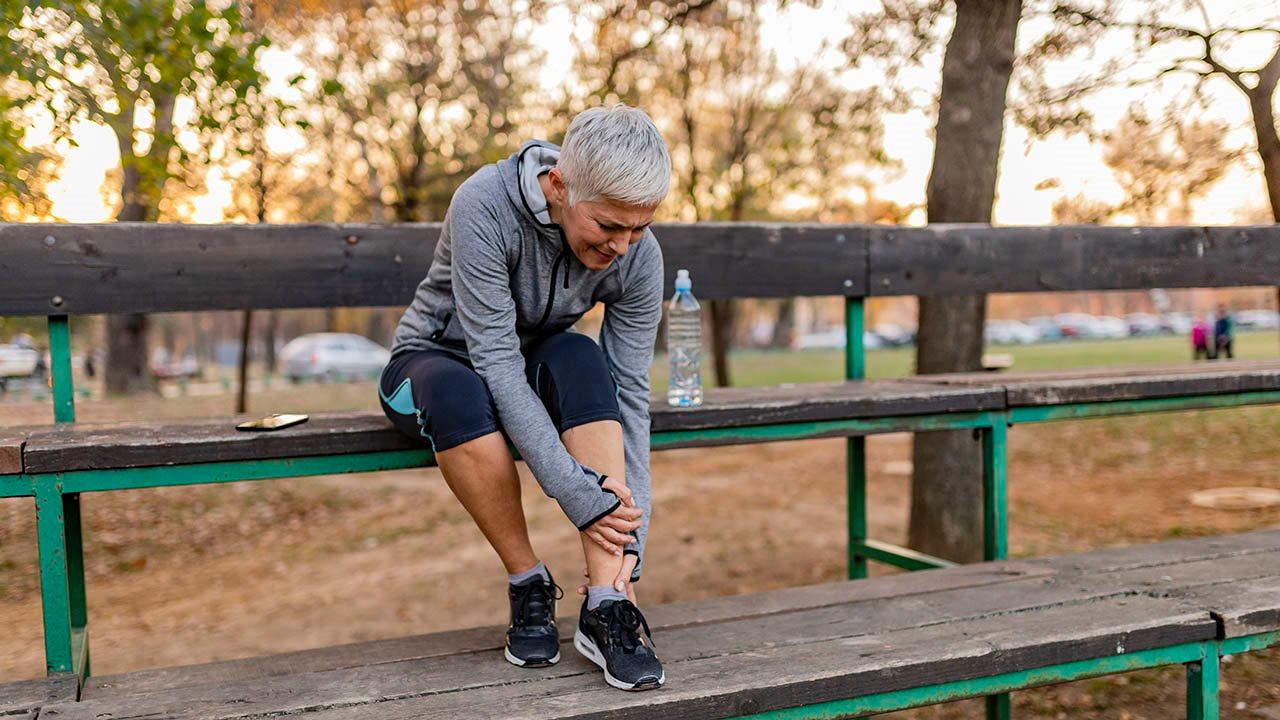Illustrasjonsfoto av en eldre dame i treningsklær med grått hår. Hun sitter på en tribune og holder seg på leggen/ankelen, noe som tyder på smerter.