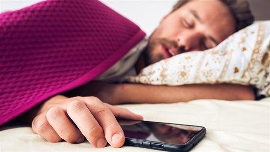 Illustrasjonsfoto: Mann som sover på siden i sengen, med hånden over en smarttelefon.