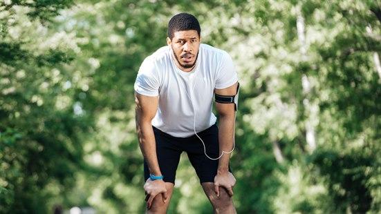 Illustrasjonsfoto: Sliten jogger i hvit t-skjorte og svarte shorts  står fremoverbøyd og lener seg mot knærne. I bakgrunnen ser vi grønt bladverk fra trær.