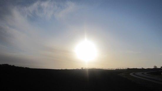 Fotoet viser vintersolen som står lavt på en blå himmel. Landskapet er ganske flatt. På høyre nedre hjørne av bildet er det en landevei som svinger mot høyre. 