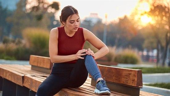 Illustrasjonsfoto av en kvinne i treningsklær som sitter på en trebenk i en park. Hun har den ene foten plassert på benken og tar seg på kneet.