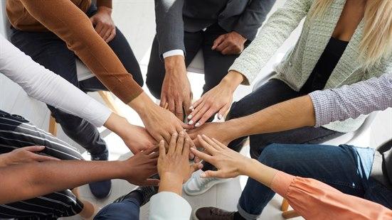Fotoet viser armene og hendene til åtte voksne personer som sitter på stoler i en liten sirkel og strekker én arm hver til midten av sirkelen, der hendene treffes. 