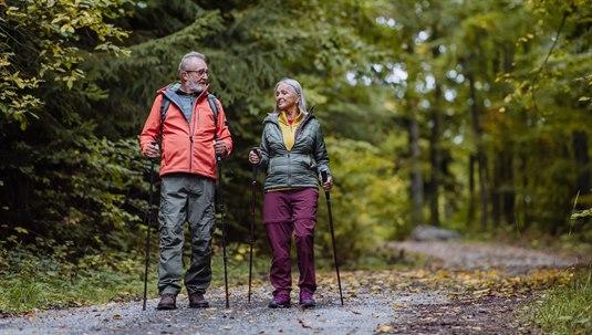 På en skogsti går et par i pensjonsalderen, utstyrt med turlær og staver. De ser ut til å ha det fint mens de går tur og nyter naturen rundt seg.