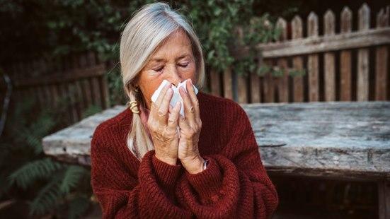 Fotoet viser en godt voksen kvinne ikledd en rød genser. Hun sitter ute ved et solid hagebord med øyne lukket og pusser nesen med et lommetørkle.