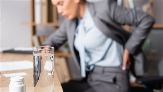 Kvinne som sitter ved en kontorpult og tar seg til ryggen, angir smerter i bekkenet på venstre side. Glass med smertestillende medisiner foran henne på pulten