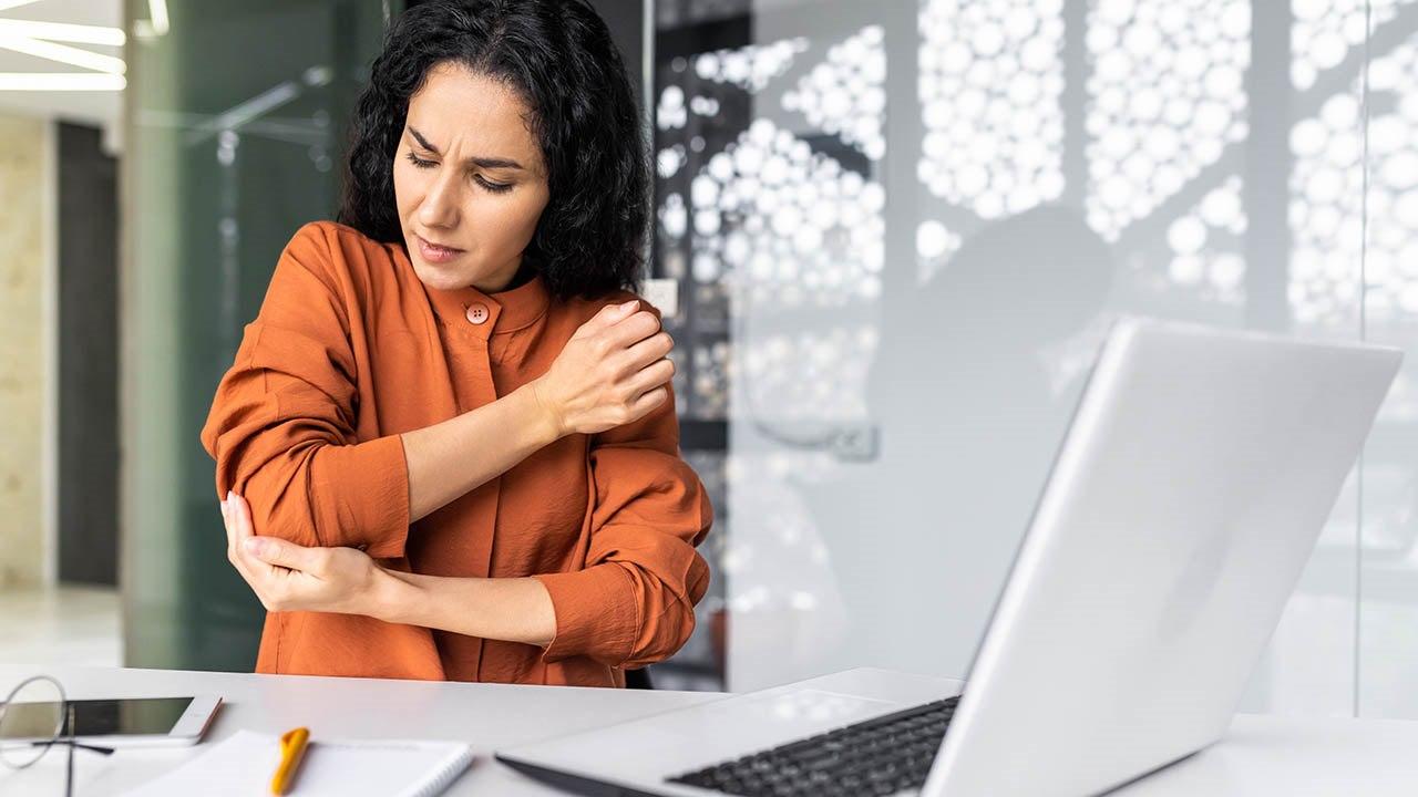 Illustrasjonsfoto av kvinne med mørkt hår og oransje skjorte som holder seg på albuen og viser smerte. Hun ved arbeidspulten sin.