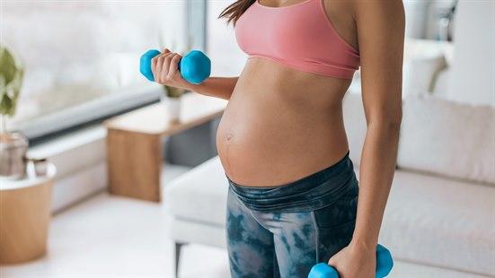 Illustrasjonsfoto: Utsnitt av kroppen til gravid dame fra lårene og til over brystene. Hun har rosa treningstopp og grå-sorte treningsbukser. Magen er bar og hun holder to manualer i hendene.