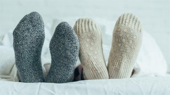 Illustrasjonsfoto: To par føtter som stikker ut under dynekanten. Dynen er hvit. To av ullsokkene er hvite, to er grå.