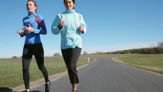 Fotoet viser to kvinner i trettiårene på joggetur. De springer på en smal asfaltvei i strålende solskinn. Fotoet er tatt forfra.
