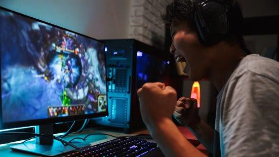 Illustrasjonsfoto: Ung gamer med hodetelefoner sitter foran en skjerm og et tastatur og jubler.