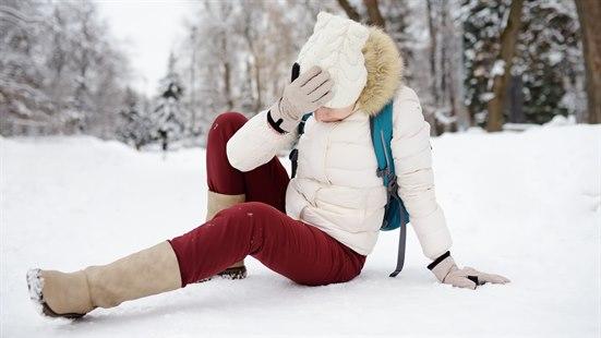 Kvinne sitter på baken i snøen og tar seg til hodet. I bakgrunnen er snødekte trær.