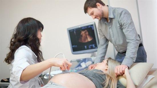 Illustrasjonsfotoet viser en gravid kvinne i følge med sin mann som undersøkes med ultralyd. Ultralydoperatøren sitter med hodet vendt mot ultralydskjermen og den gravide ligger på en benk