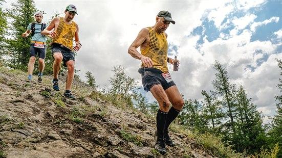 Illustrasjosfoto: Tre menn deltar i løpskonkurranse i krevende terreng. Løper med vannflasker i hendene, ned bratt, steinete bakke.