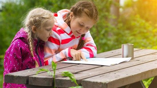 Illustrasjonsfoto av to jenter som sitter på en benk i naturen og studerer et kart. 