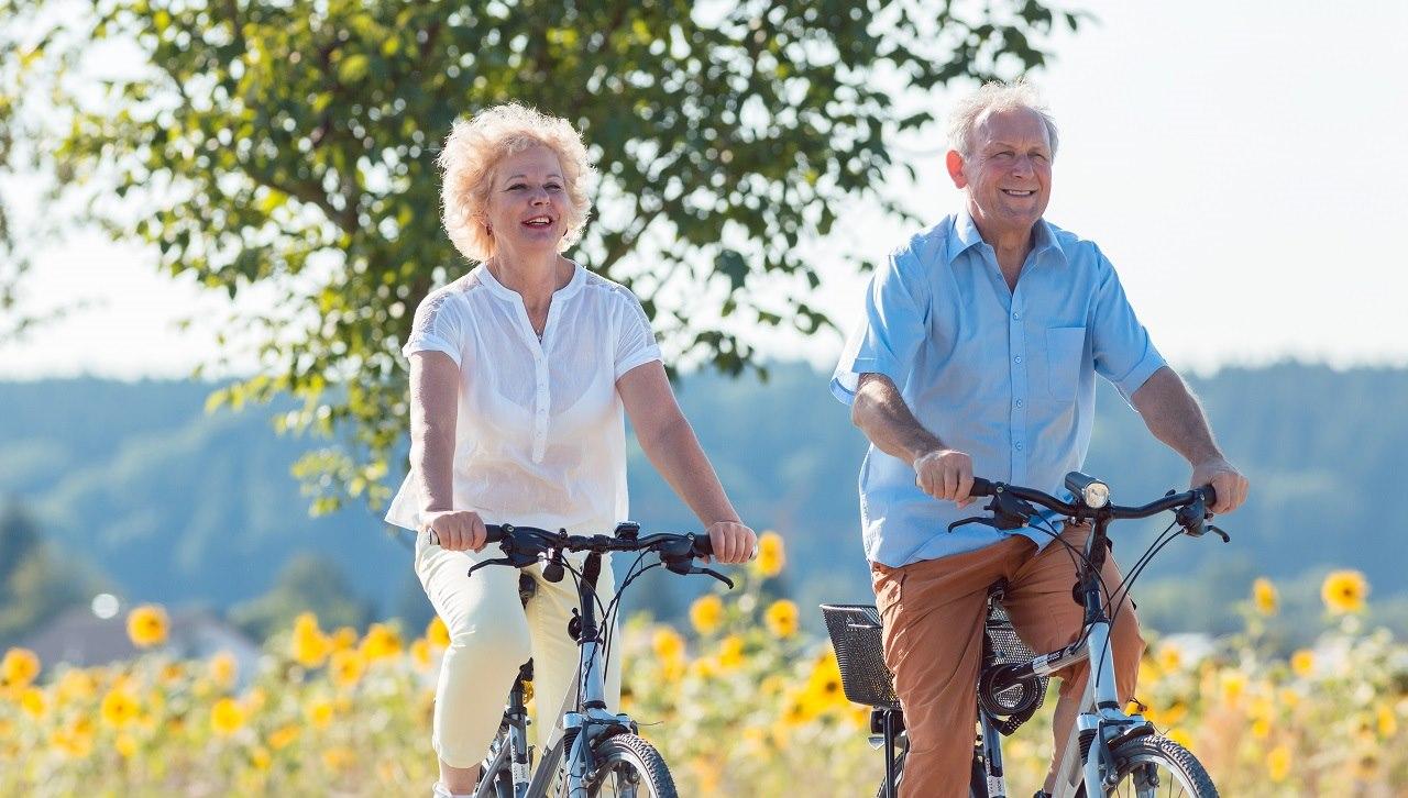 Illustrasjonsfoto av en mann og kvinne som sykler på en sti med en blomstereng og trær i bakgrunnen.