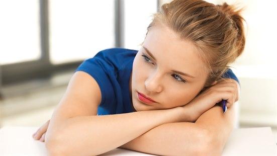 Illustrasjonsfoto av en ung, tankefull kvinne sitter lent over en pult, med hodet hvilende på overarmene. 