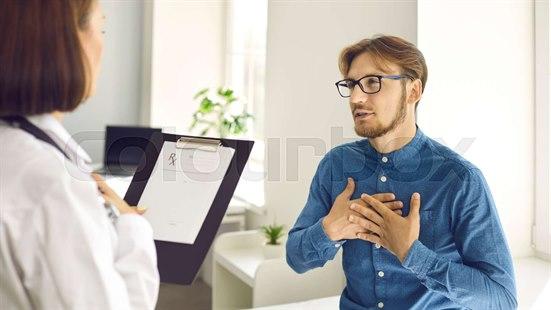 Illustrasjonsfoto av en ung mann med briller og blå dongeriskjorte sitter på en benk hos legen. Han holder hendene foran brystet. Vi ser ryggen og hpret til legen, som holder en notatbok foran seg.