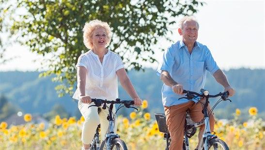 Illustrasjonsfoto av en mann og kvinne som sykler på en sti med en blomstereng og trær i bakgrunnen.