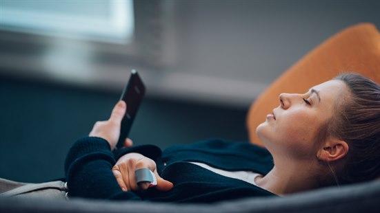 Dame som ligger på sofaen og slapper av. Holder smarttelefon i en hånd, og har et fingerbøl på en finger.