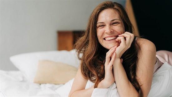 Illustrasjonsfoto av en ung, smilende kvinne med langt brunt hår og fregner som ligger på magen i sengen, hviler hodet i hendene og ser mot kameraet.