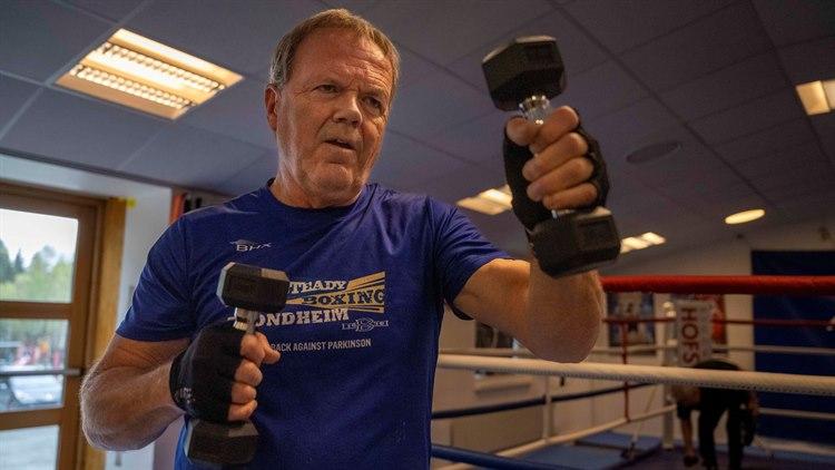 Mann i blå t-skjorte trener med en manual i hver hånd. Han er i em boksesal.