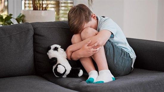 På sofaen sitter et lite barn sammen med bamsen sin, tilsynelatende lei seg og trist, med hodet gjemt mellom beina.