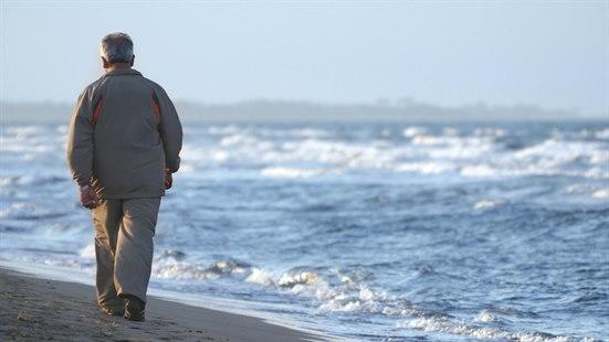 Illustrasjonsfoto av en eldre mann som går langs strandkanten. I bakgrunnen sees sjøen, fjell og noen bygninger langt borte ved horisonten.