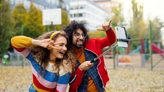 En dame og en mann som poserer foran et mobilkamera festet på en selfiestang.