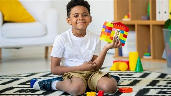Illustrasjonsfoto: Gutt som sitter på et svart og hvitt teppe i en stue. Beina hans er i W-stilling, med leggene på utsiden av lårene. Han holder et hus han har bygget i lego i den ene hånden og smiler til kameraet.
