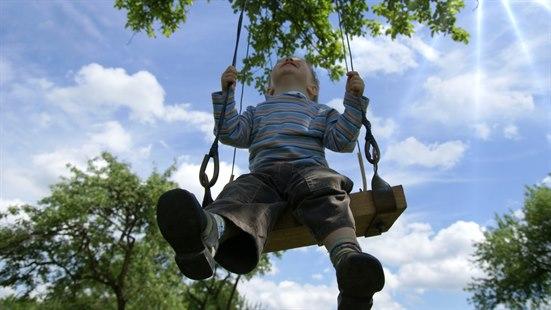 Illustrasjonsfotoet viser en liten gutt som gynger på en huske. Himmelen er blå, og solen skinner.