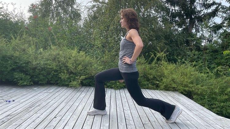 Kvinne i svart treningstights og grå singlett tøyer hofteleddsbøyer ute på grå platting. Trær og busker i bakgrunnen.